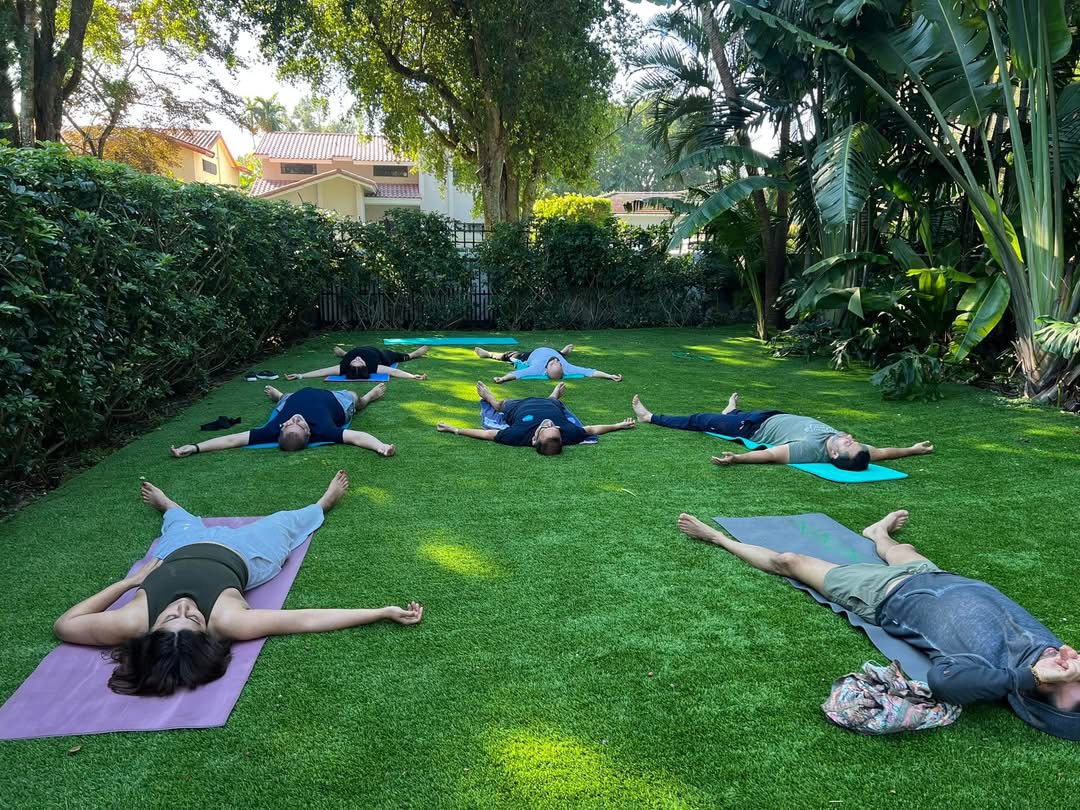 A group of people resting on colorful yoga mats in a quiet outdoor session on a grassy lawn surrounded by trees
