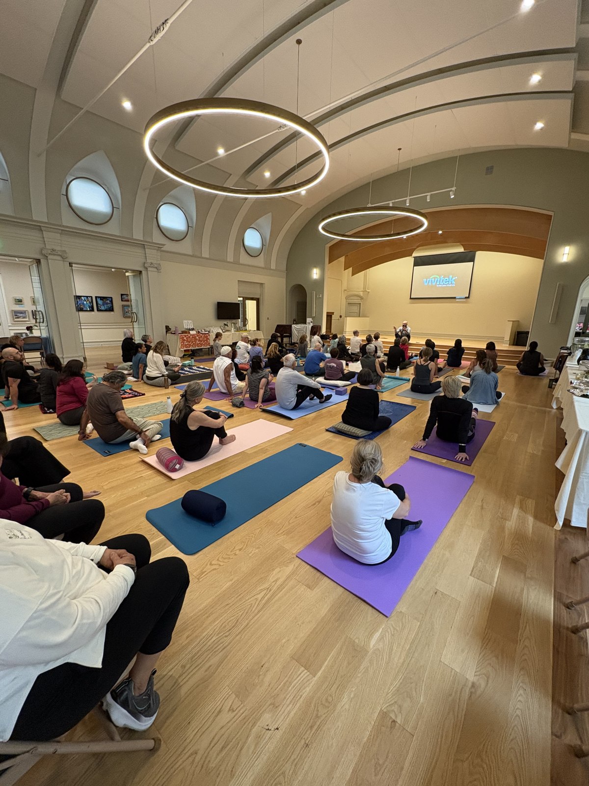 Dr. Saira Hussain leading a community wellness workshop from a small stage, with about thirty participants seated on colorful yoga mats in a bright hall