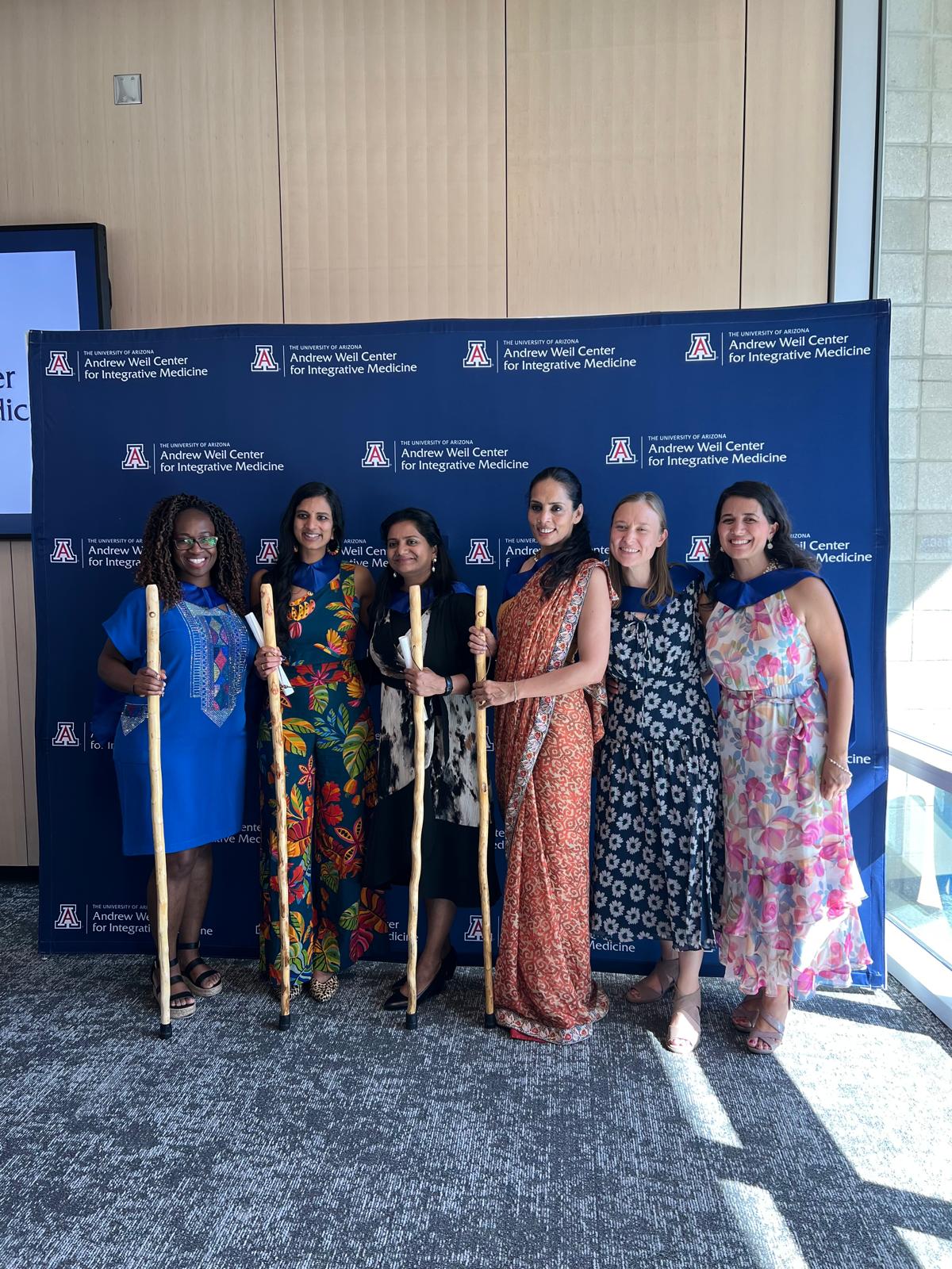 Dr. Saira Hussain with five fellow graduates of the Andrew Weil Center for Integrative Medicine, holding wooden walking staffs in front of a branded backdrop