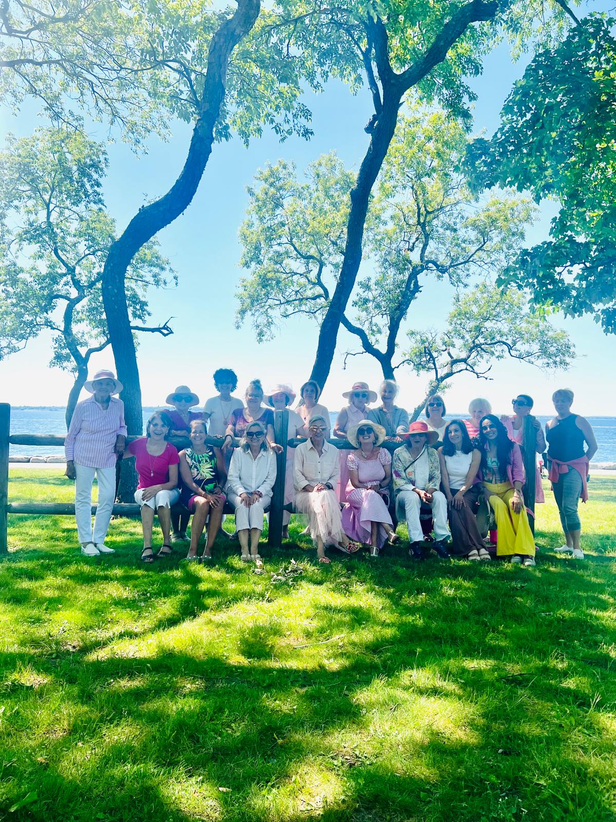 About twenty women in summer dresses and sun hats gathered along a wooden fence above the water during the RIHWC spring wellness walk