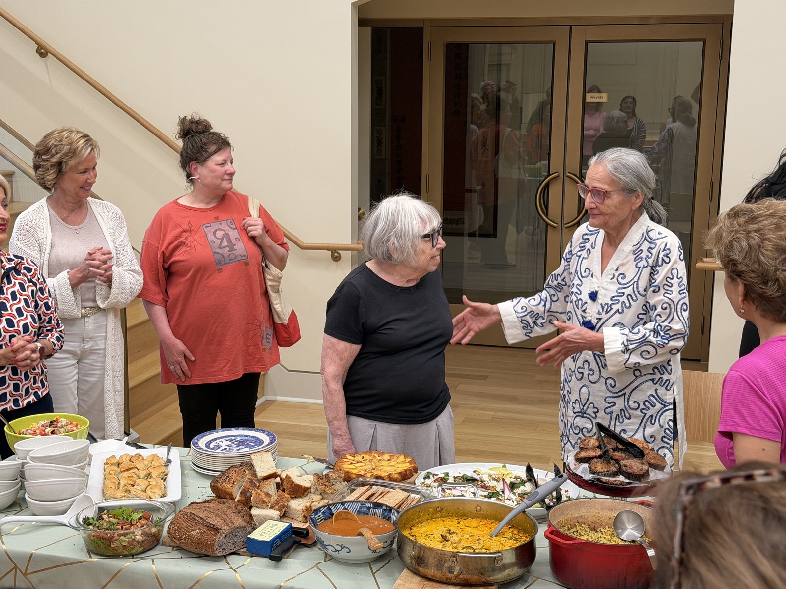 A group of women gathered around a long table covered with salads, bread, pastries and curry, talking together in a bright room.