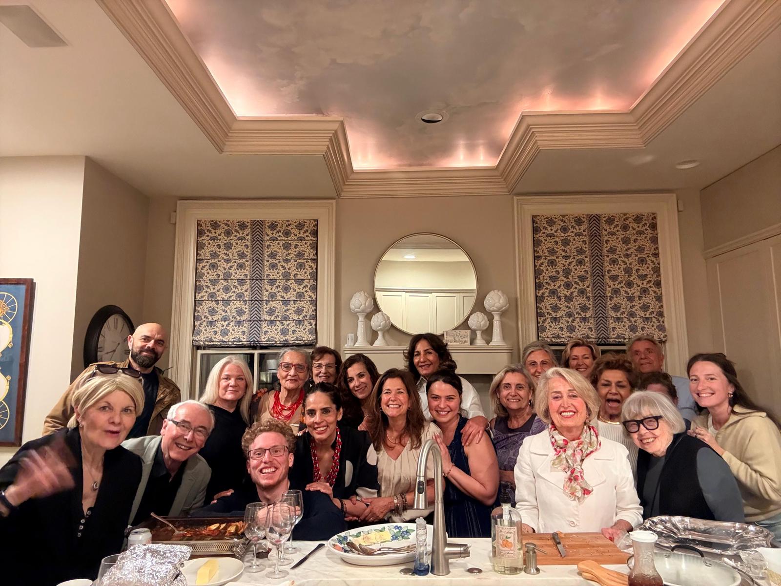 A diverse group of about twenty people of all ages smiling together behind a kitchen island filled with shared dishes during a community gathering