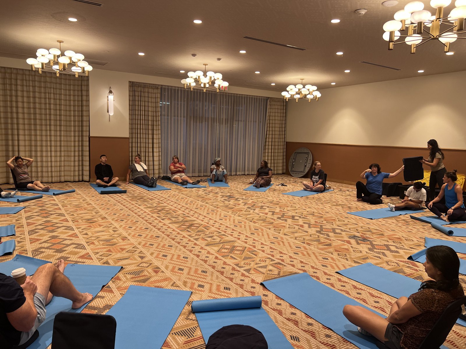 About fifteen people sitting in a wide circle on blue yoga mats during a wellness session, with an instructor holding a prop at the front