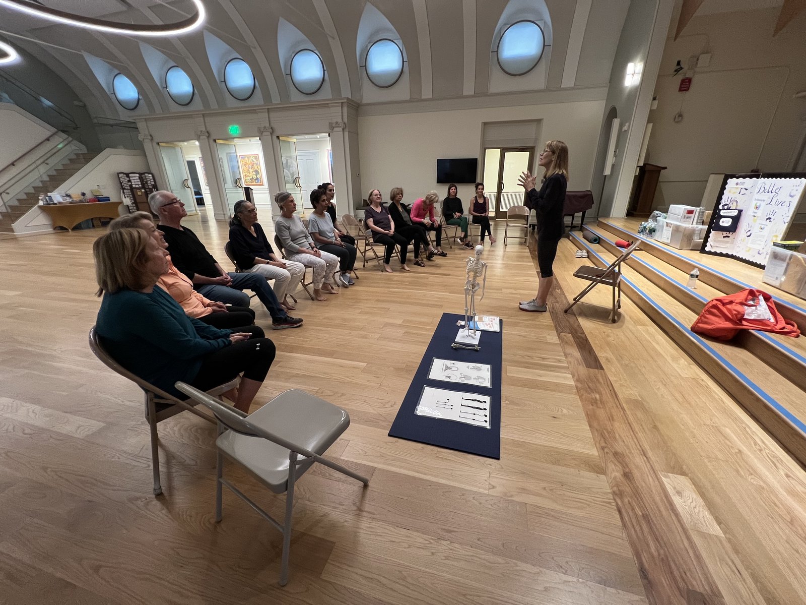 A group of adults seated in a semi-circle in a bright hall listening to a presenter, with a skeleton model and diagrams on a mat nearby.