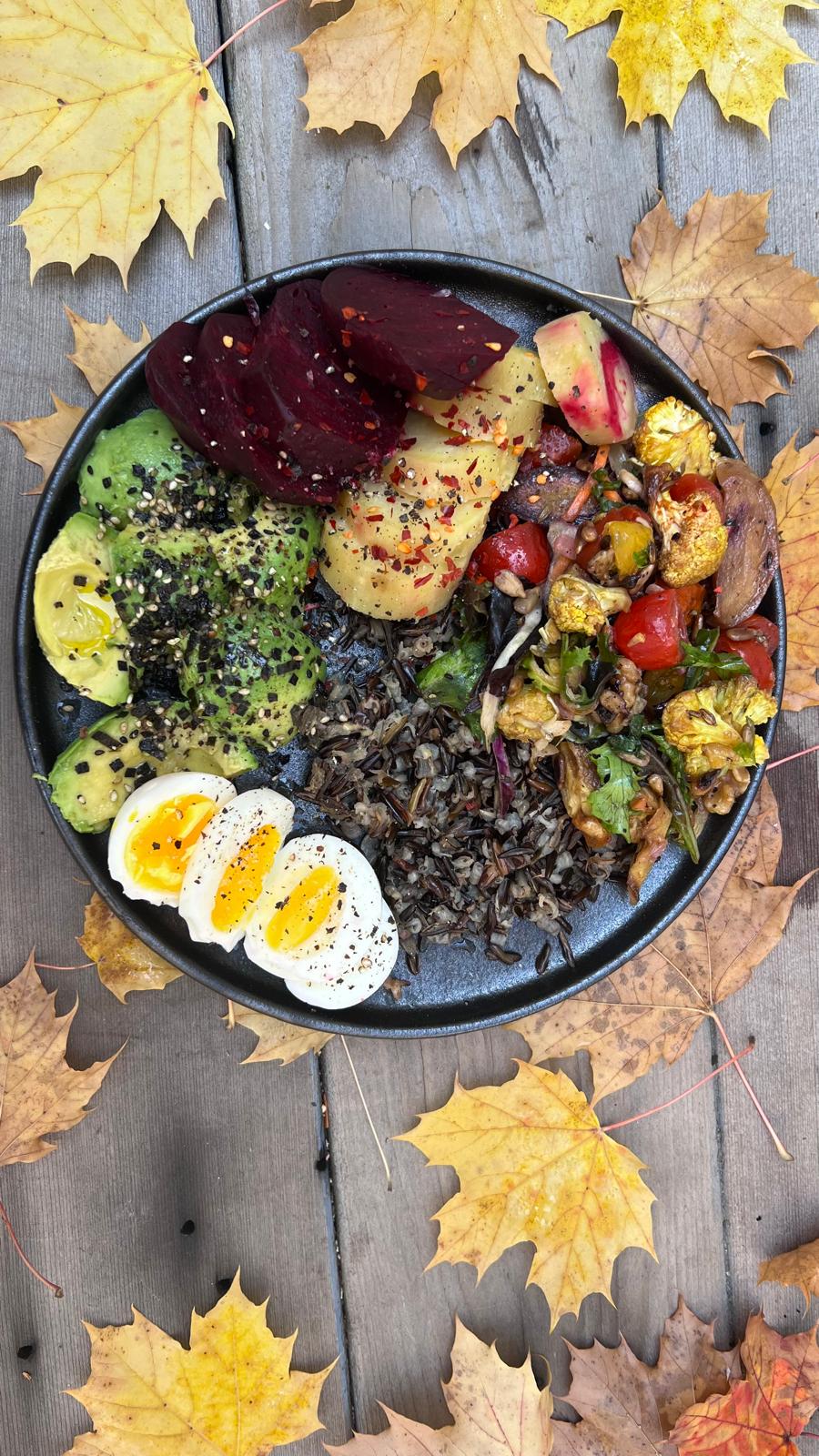 A colorful plant-forward bowl with avocado, roasted vegetables, beets, soft-boiled eggs and wild rice on a wooden table with autumn leaves