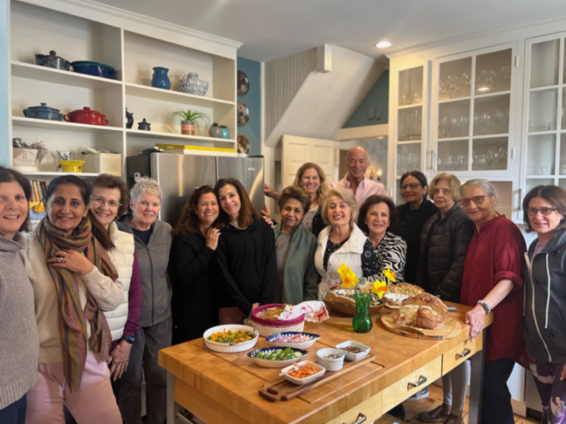 A smiling group gathered around a wooden island in a bright kitchen, with bowls of food and colorful ceramic dishes on the shelves behind them.