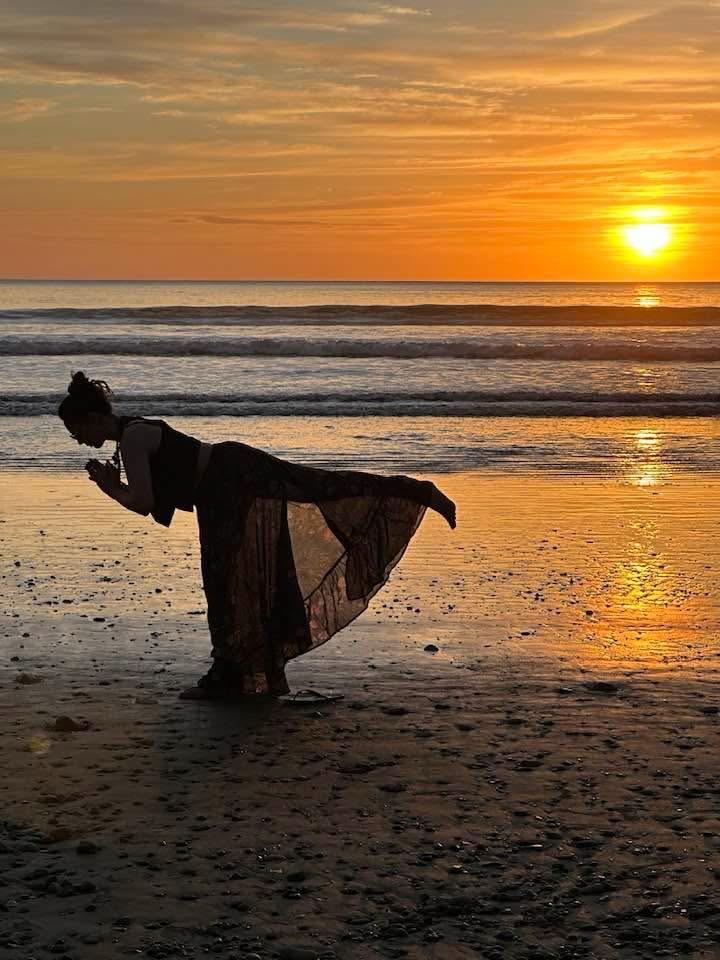Silhouette of a woman holding a balanced yoga pose on a beach at sunset, with ocean waves and a glowing orange sky behind her