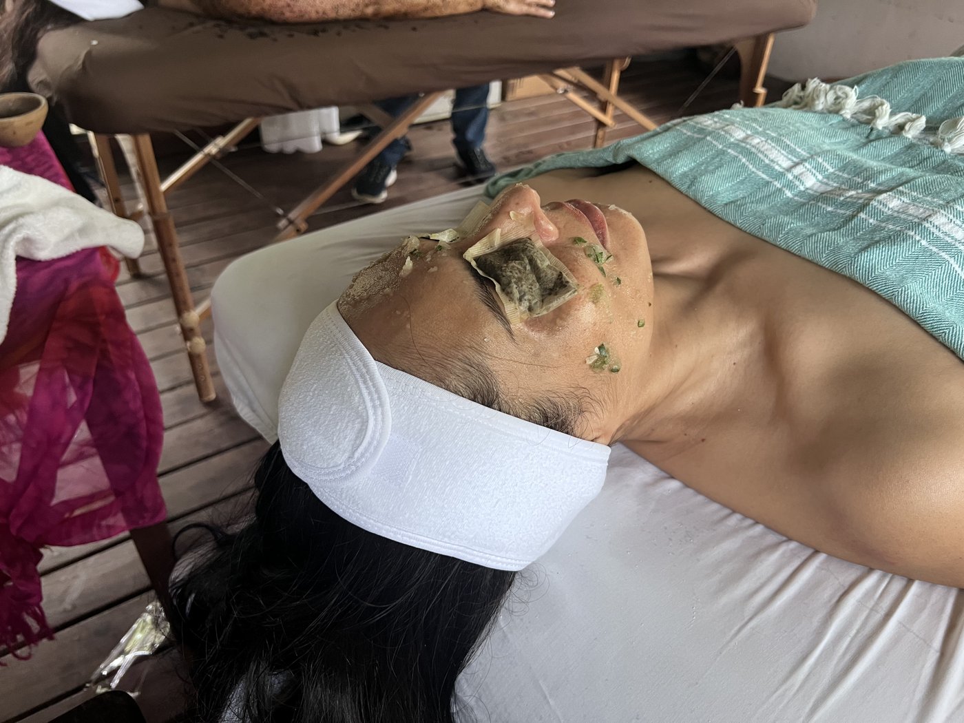 A person lying on a white treatment table outdoors with herbal tea bags over their eyes and green botanicals on their cheeks during a relaxing spa treatment