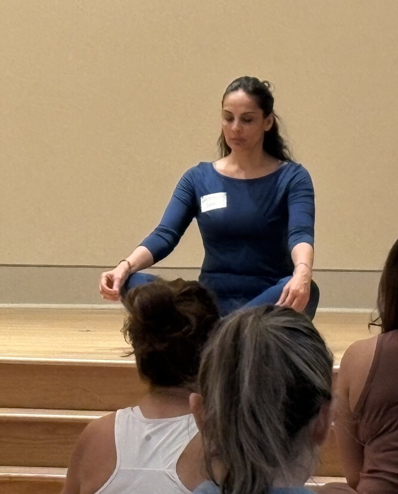 An instructor seated cross-legged on a wooden stage leading a group meditation session with her eyes closed, as participants sit facing her